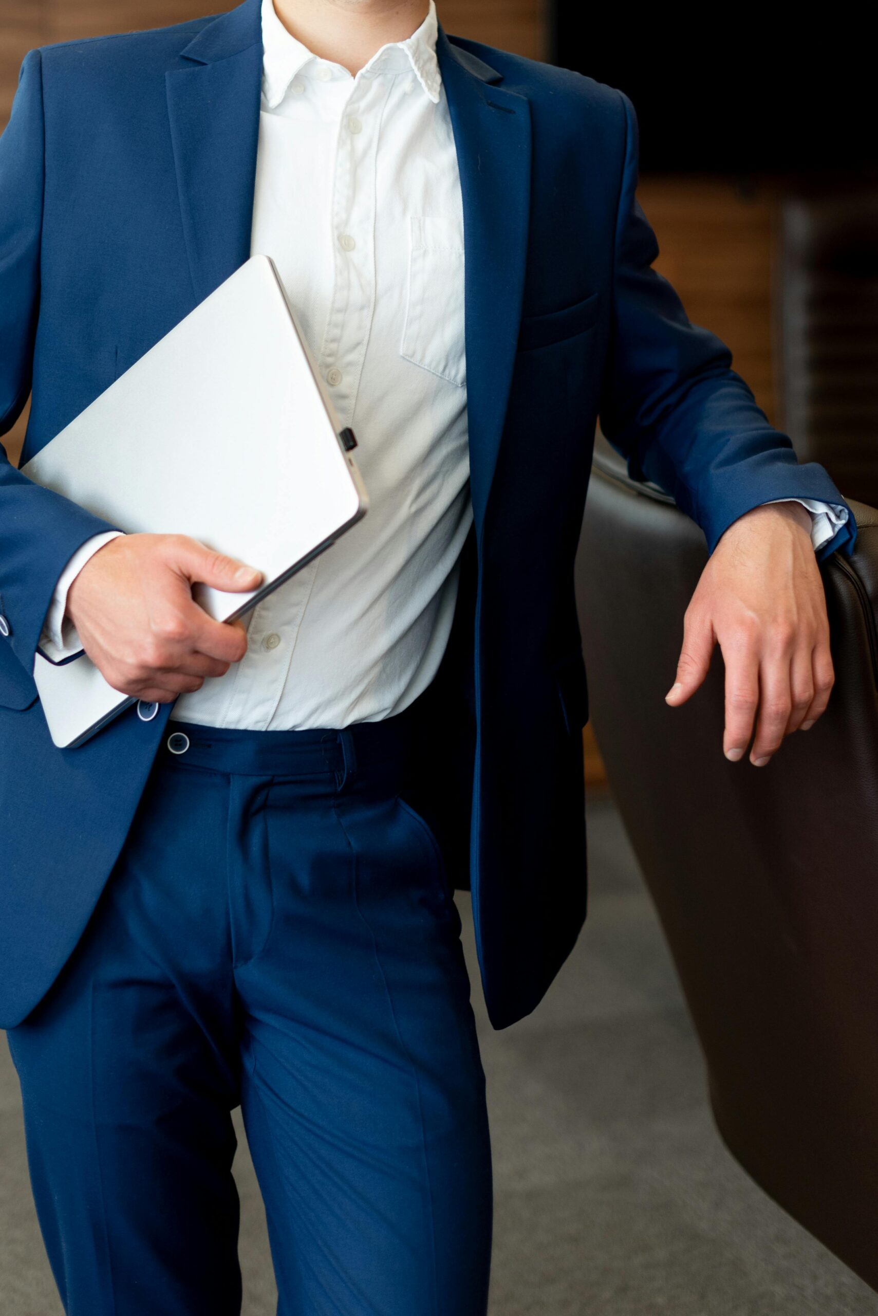 Business professional in blue suit holding a laptop, leaning casually in office environment.